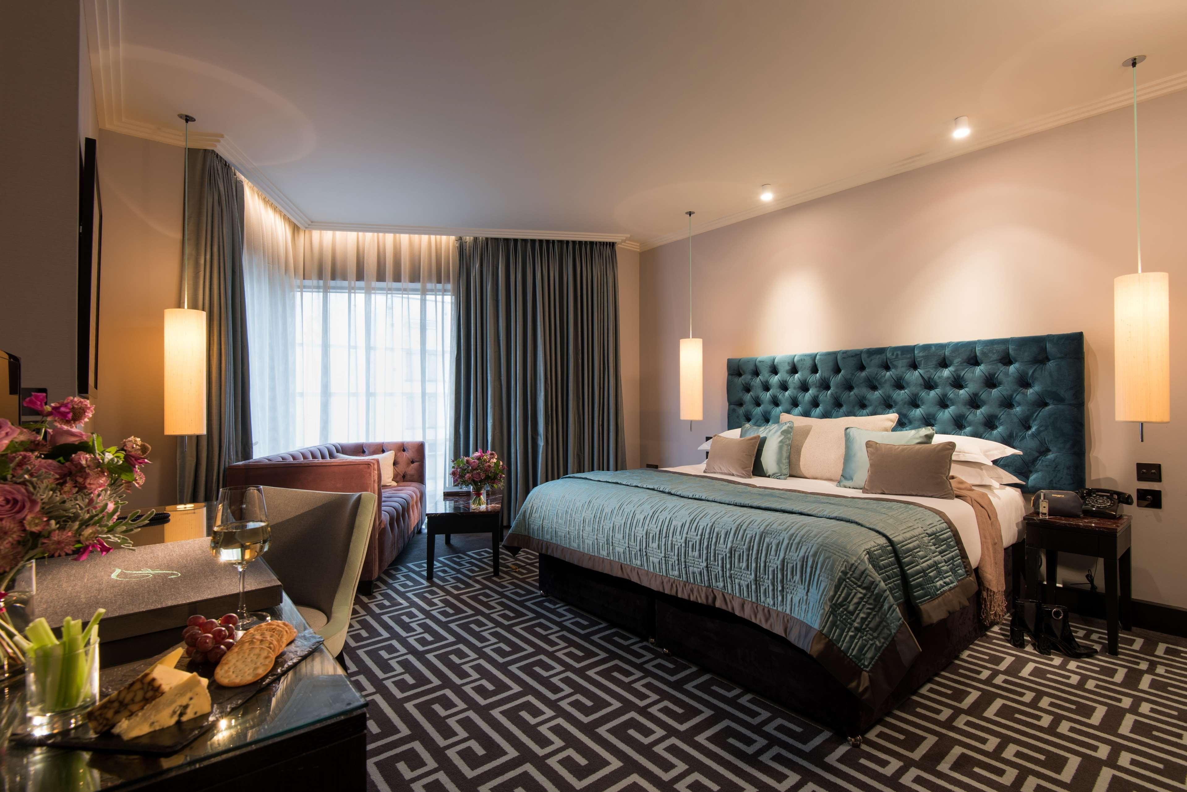 The Fitzwilliam Hotel Belfast Kültér fotó The photo depicts a stylish hotel room. It features a large bed with a tufted teal headboard, flanked by two wall sconces providing soft lighting. The bedding is neatly arranged with accent pillows. To one side, there is a small seating area with a c