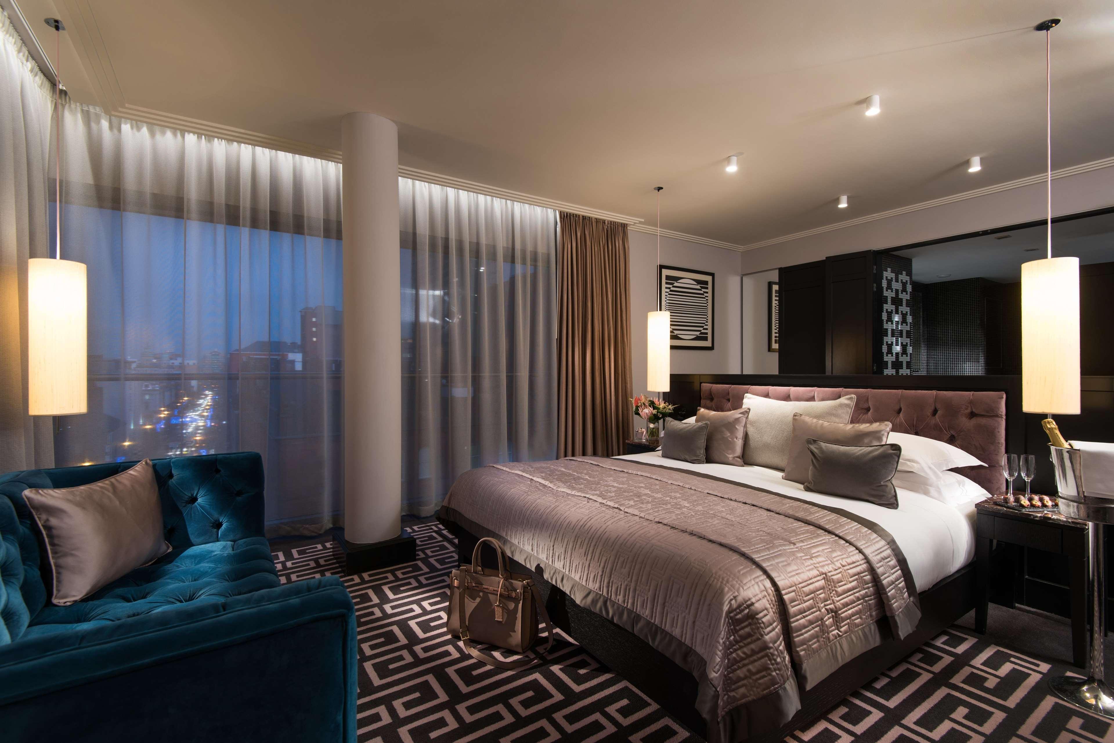 The Fitzwilliam Hotel Belfast Kültér fotó The photo shows a luxurious hotel room with a modern design. It features a large bed with decorative pillows and a stylish bedspread, complemented by bedside lamps. To one side, there is a plush turquoise sofa and a coffee table. The room has floor-t