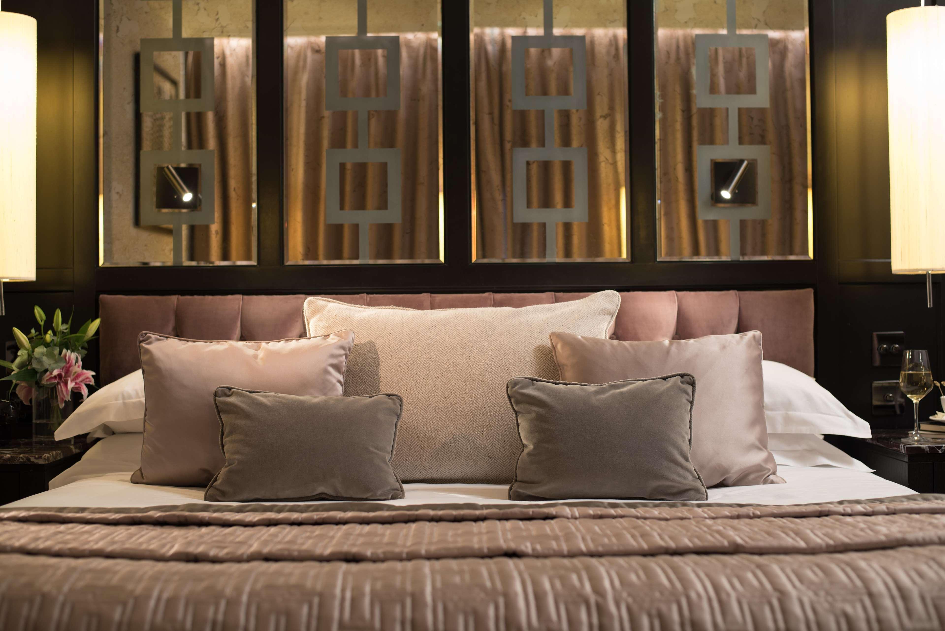 The Fitzwilliam Hotel Belfast Kültér fotó The photo shows a beautifully designed bed in a stylish bedroom. The bed is adorned with a soft, plush headboard and layered with various decorative pillows in shades of beige and gray. The bedding appears luxurious, with a textured blanket. Behind t