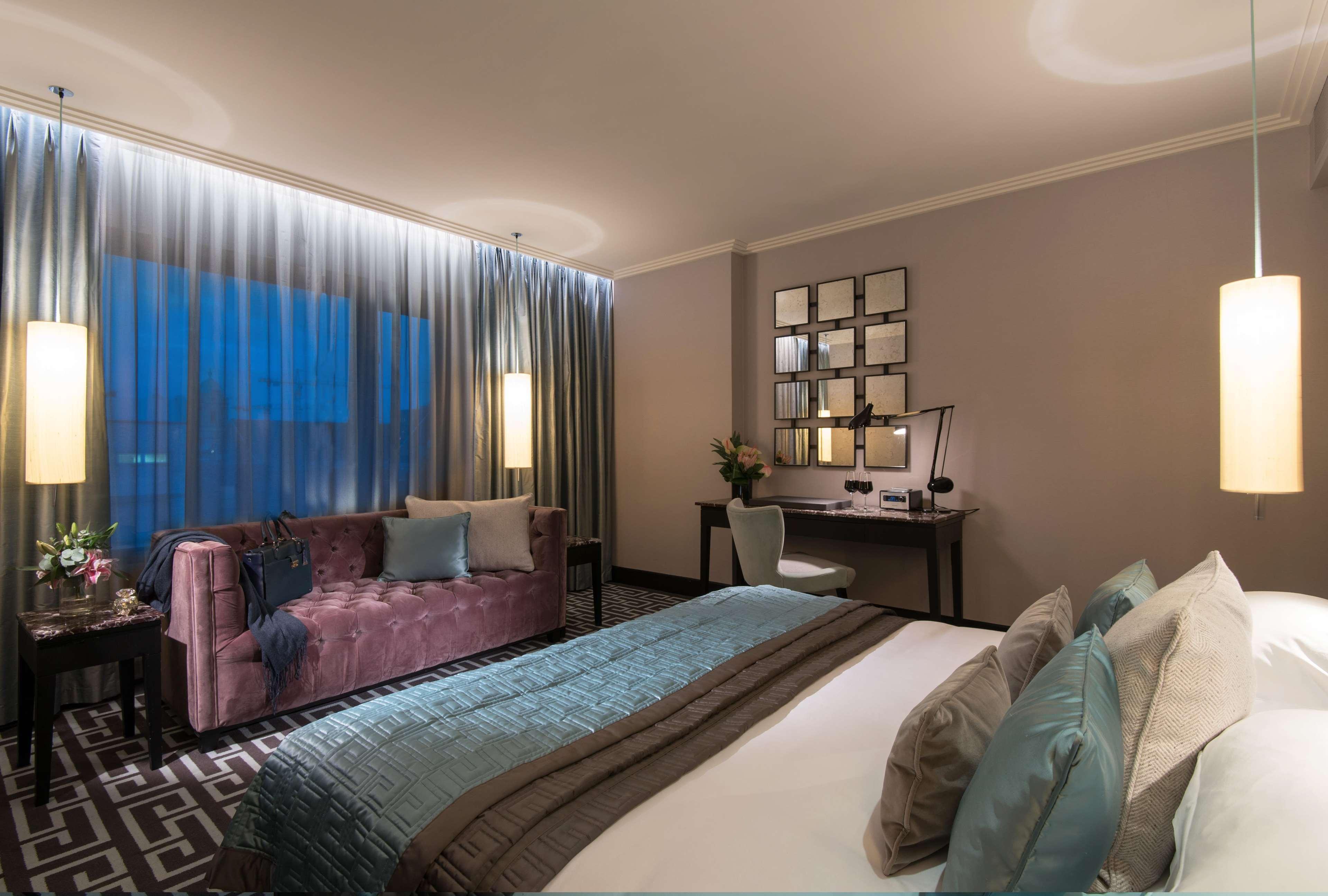 The Fitzwilliam Hotel Belfast Kültér fotó The photo depicts a modern hotel room featuring a spacious layout. There is a large bed with decorative pillows in the foreground, covered with a stylish bedspread. To one side, a plush purple sofa adds a pop of color and comfort. A small desk and ch