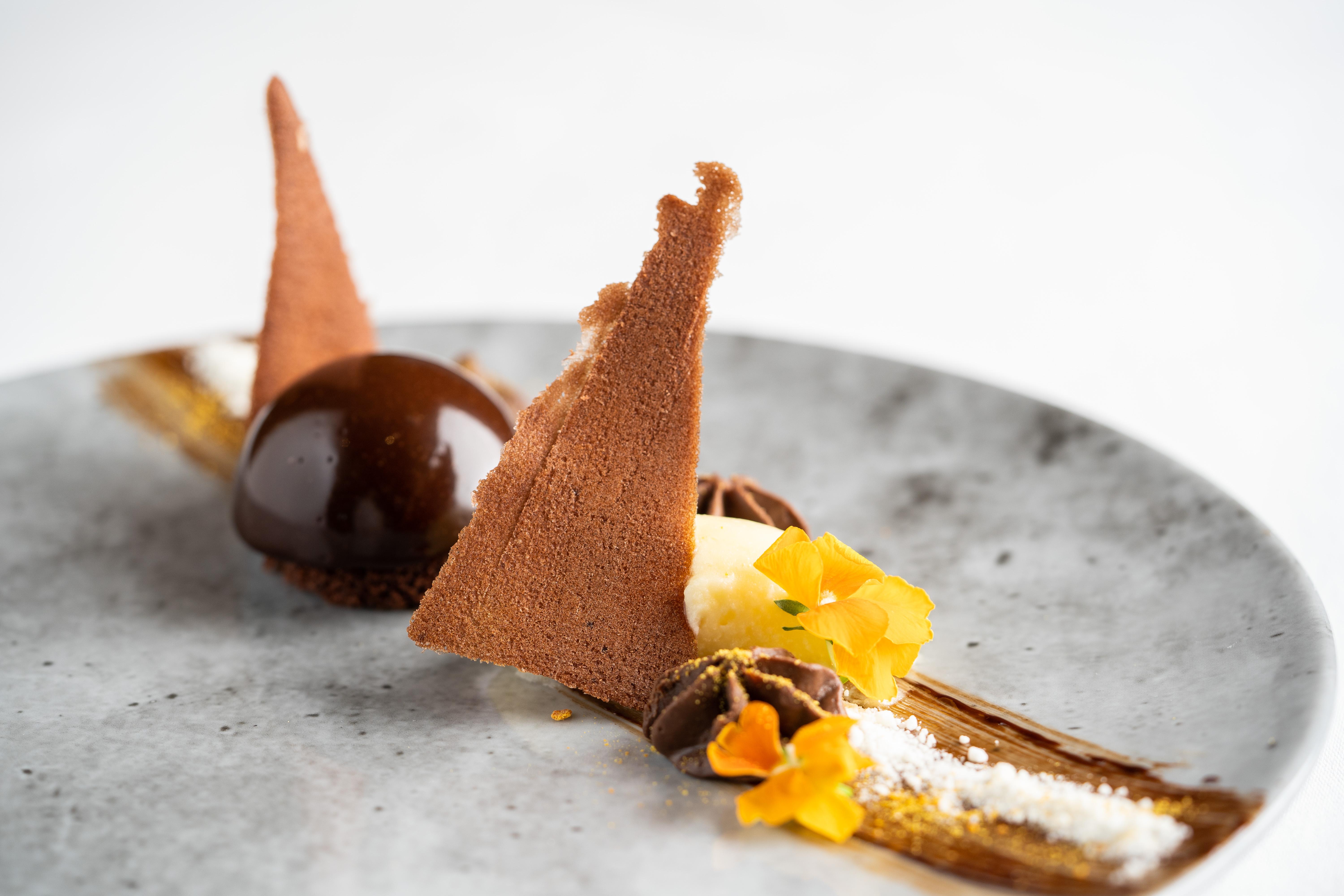 The Fitzwilliam Hotel Belfast Kültér fotó The photo displays an artfully plated dessert. It features a glossy chocolate sphere, accompanied by various chocolate components, including triangular chocolate tuile pieces that resemble sails. The plate is adorned with delicate garnishes such as y