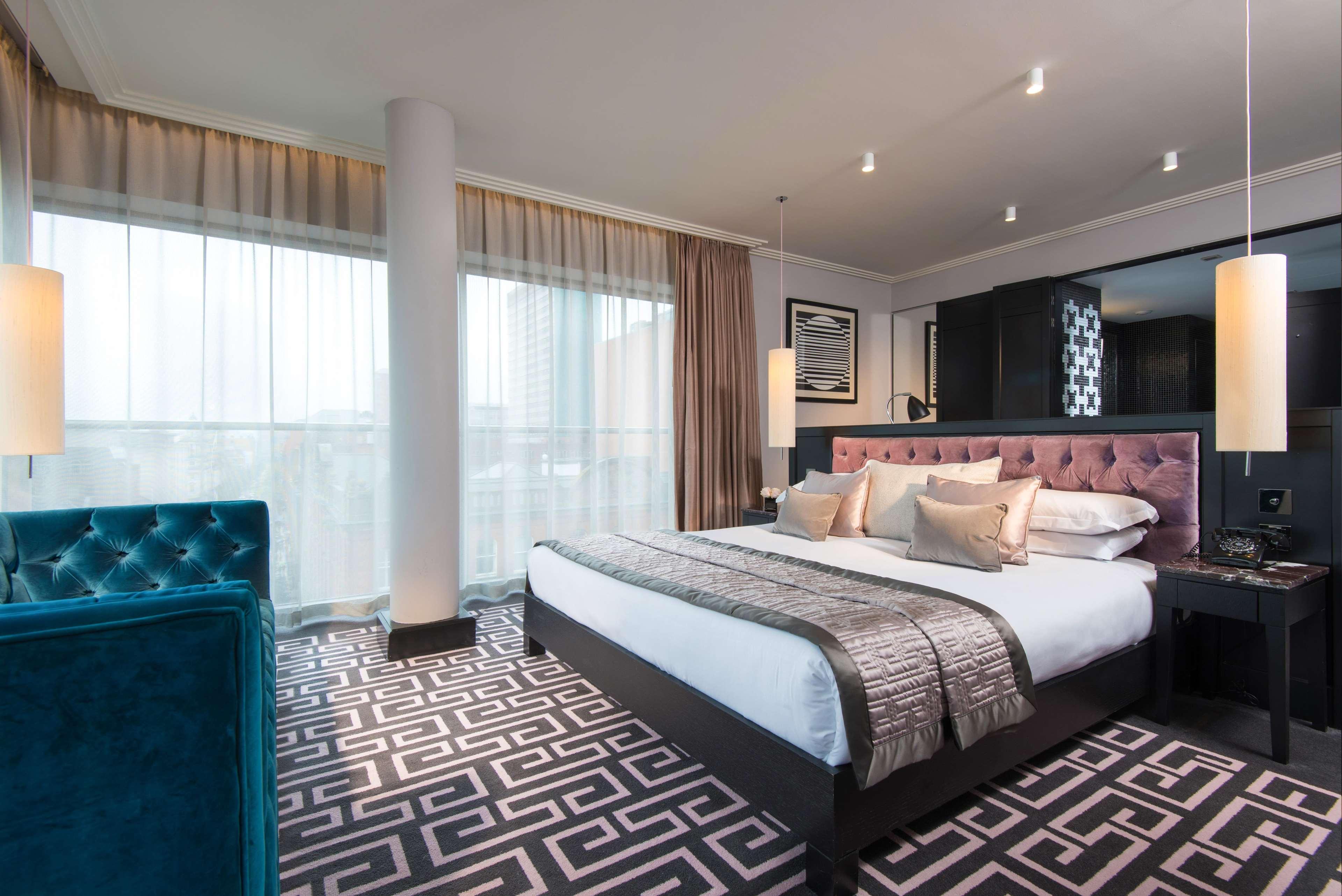The Fitzwilliam Hotel Belfast Kültér fotó The photo showcases a modern hotel room. It features a large bed with plush pillows and a stylish headboard. The room has large windows with sheer curtains, allowing natural light to filter in. The floor is covered with a patterned carpet that contra