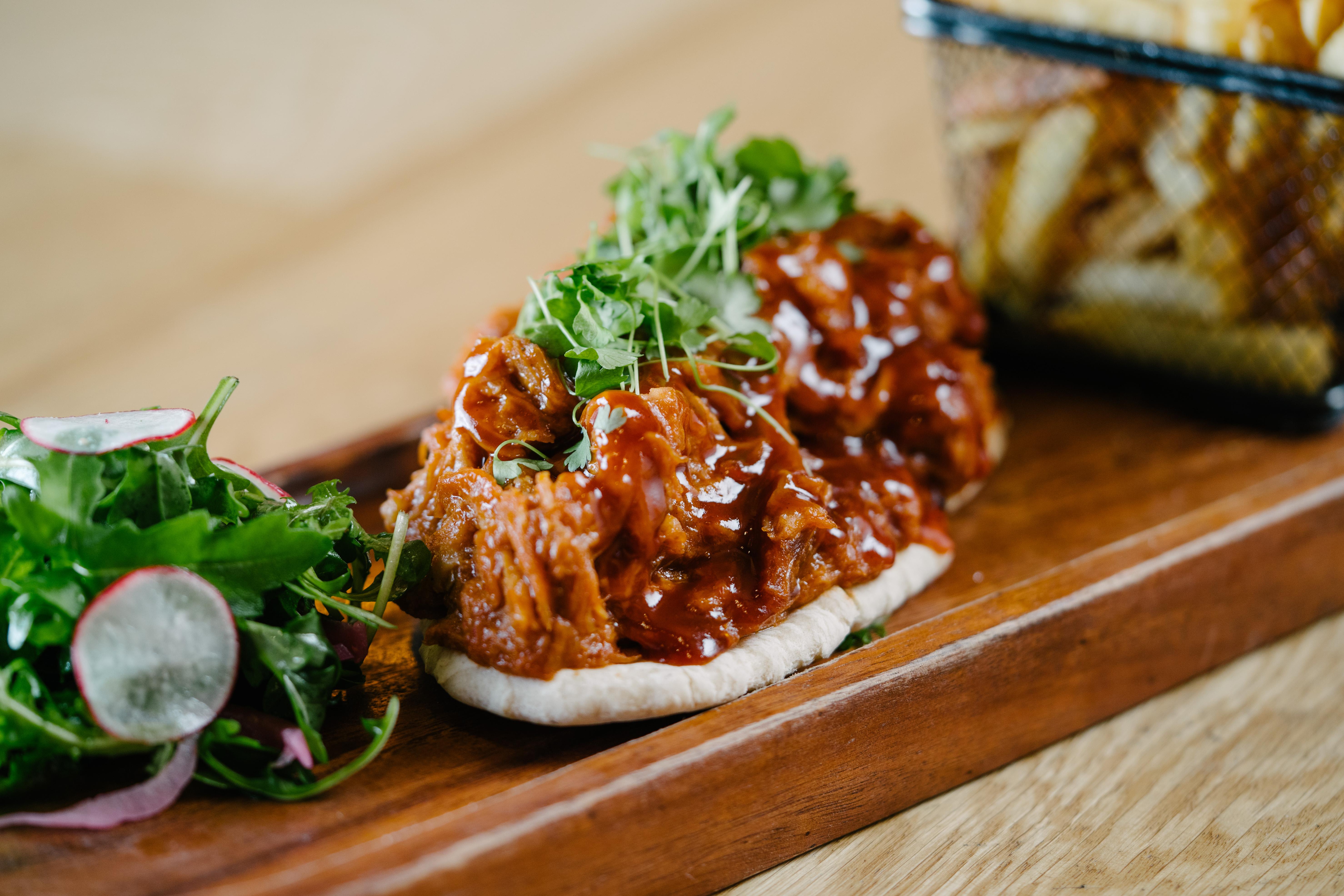 The Fitzwilliam Hotel Belfast Kültér fotó The photo shows a wooden platter featuring two pieces of bread topped with pulled meat covered in a rich barbecue sauce. The dish is garnished with fresh greens, including sprouts. On the side, there is a small serving of mixed salad with radishes an